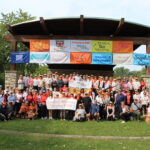 A large group of people stand on a stage and look towards the camera. The stage has a variety of colorful banners hanging at the top. The front row of people holds a sign reading "Chicagoland Walk and Roll to Cure FSHD"