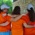 Three women wearing orange shirts and grey baseball hats stand with their arms wrapped around each other in a hug.
