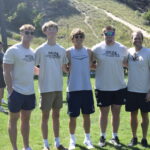 5 men looking towards the camera stand with their arms around each other.