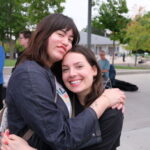 2 women smile at the camera as they hug. The woman on the left sits in a power chair.
