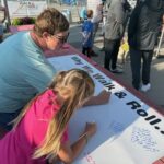 An older woman and a girl write with markers on a large white banner that says "Why We Walk & Roll"