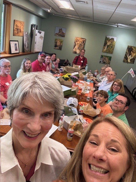 A selfie with two women at the front and multiple individuals sitting behind them looking at the camera while eating lunch.