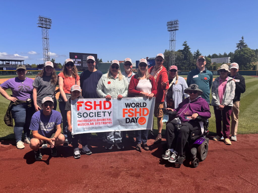 A group of individuals stand and sit around a sign for the Pacific Northwest Chapter. Some individuals are in power chairs and others use assistive devices like walkers and canes. Many of them wear baseball hats.