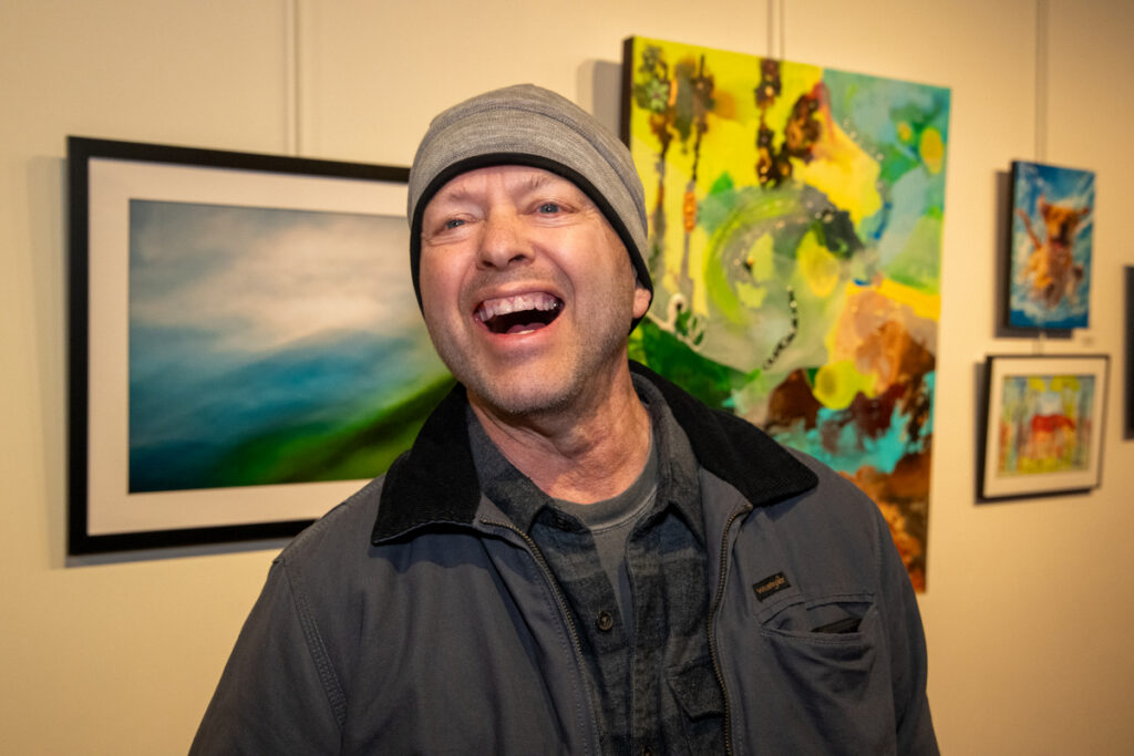 Photo of Rick, a white man wearing a grey beanie and a dark winter jacket. He stands in front of photographs and paintings in an art gallery. His mouth is open in a joyous expression.