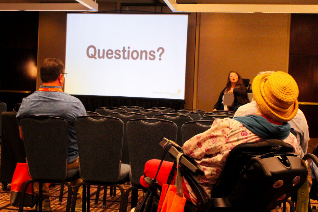 Audience members, some in chairs and some in powerchairs sit and listen to Marisa Spain end her presentation. Marisa sits in her powerchair at the back of the room next to a large screen reading "Questions."