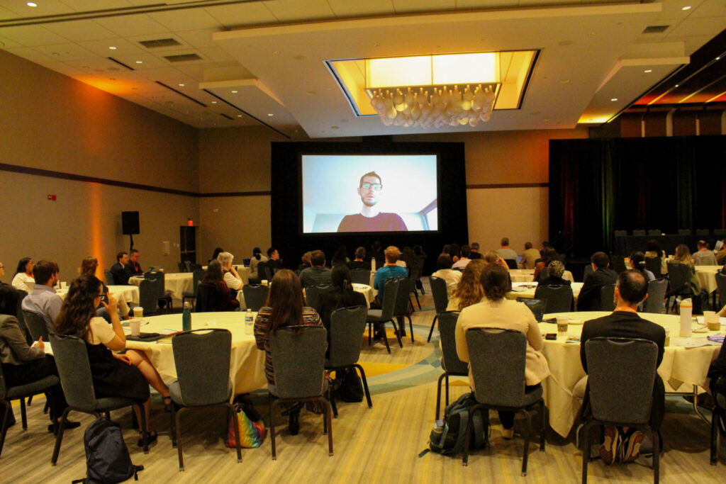 A large group of scientists and researchers sit at conference tables listening to a patient testimonial projected onto a large screen.
