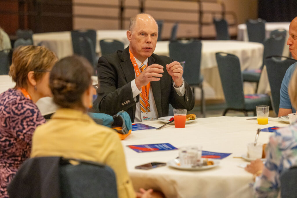 A middle aged white man sits a table talking intently with fellow conference attendees.