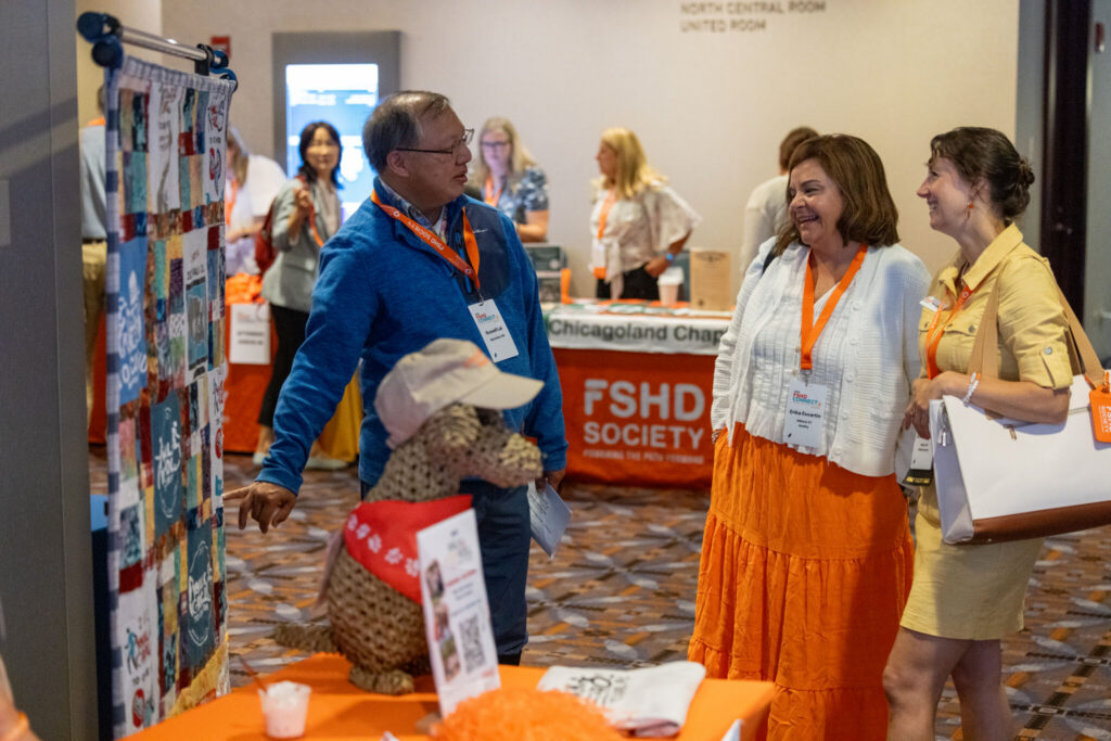A tall Asian man in a blue sweaters stands talking to two women about a quilt made up of Walk & Roll fundraiser imagery.