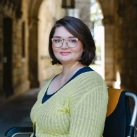 Marisa Spain, a white woman in her 20s-30s, sits in her wheelchair turned to face the camera, smiling. She wears a light yellow v-neck sweater over a darker blue v-neck shirt. Her hair is cut in a bob just past her chin. She wears peach colored glasses, which frame her blue eyes.
