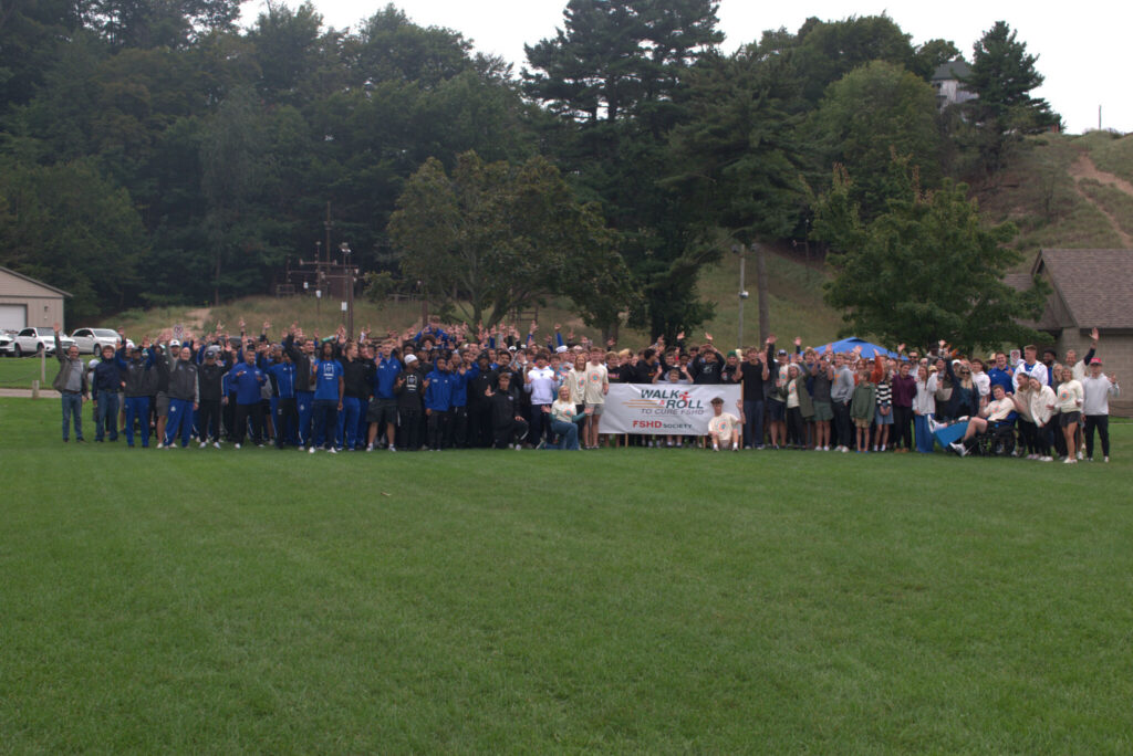 Image shows over 200 people including the entire Grand Valley State University football team dressed in royal blue track suits gathering around the white Walk and Roll to Cure FSHD banner. The photo is taken at a distance; the foreground is a large, grassy expanse and the background is filled with trees and a gray sky that was rainy only an hour before the event.