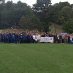 Image shows over 200 people including the entire Grand Valley State University football team dressed in royal blue track suits gathering around the white Walk and Roll to Cure FSHD banner. The photo is taken at a distance; the foreground is a large, grassy expanse and the background is filled with trees and a gray sky that was rainy only an hour before the event.