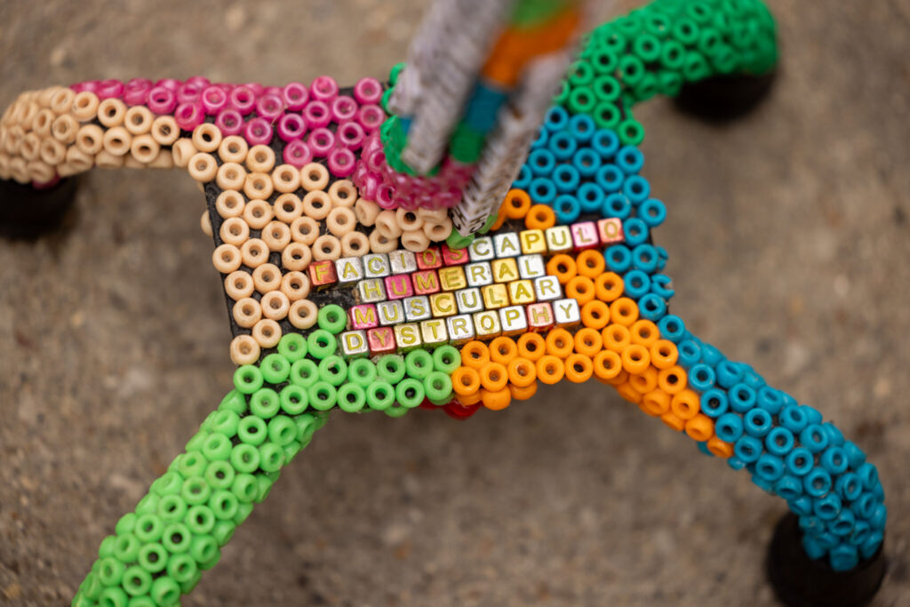 Close up photo of the base of a 4-prong cane. The cane is decorated in waves of colorful beads (light pink, bright pink, neon green, medium dark green, pumpkin orange, and a bright teal). In the center of the cane, small square beads spell out FSHD unabbreviated. These letter beads are gold, orange, and silver.