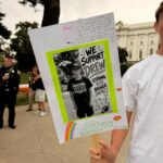 A close up of a sign made in support of Team Drew at the 2026 Sacramento Walk and Roll to Cure FSHD. Someone, out of frame, holds the poster on a stick in their right hand and they are wearing a white t-shirt. The poster is white with supportive messages (not all are legible) and a rainbow drawn on it. In the center of the poster, a bright green border frames a photo of Drew wearing a Denver Broncos shirt and hat. This photo is printed in black and white. Next to the photo, text reads: We support Drew. Strong kid. Bigger impact. Beneath these words are a cartoon football and a cartoon football strategy diagram.