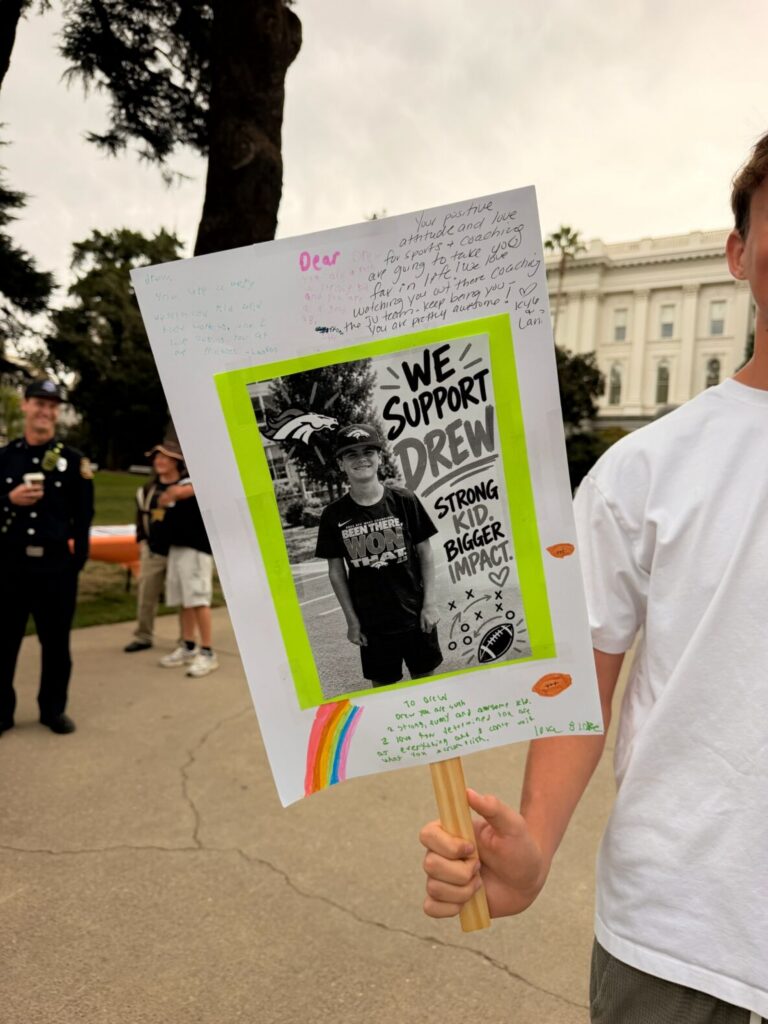 A close up of a sign made in support of Team Drew at the 2026 Sacramento Walk and Roll to Cure FSHD. Someone, out of frame, holds the poster on a stick in their right hand and they are wearing a white t-shirt.The poster is white with supportive messages (not all are legible) and a rainbow drawn on it. In the center of the poster, a bright green border frames a photo of Drew wearing a Denver Broncos shirt and hat. This photo is printed in black and white. Next to the photo, text reads: We support Drew. Strong kid. Bigger impact. Beneath these words are a cartoon football and a cartoon football strategy diagram.