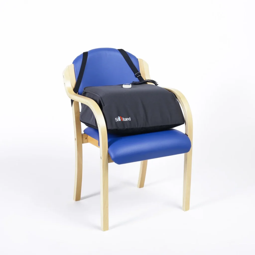 A bright, royal blue chair sits in the center of a white background. In the center of the chair, rests a large black cushion which assists individuals in getting up from the chair more easily and safely. Black straps attach the cushion to the back of the chair and provide extra stability. The cushion raises and lowers to assist individuals from a sitting to standing position more safely and easily.