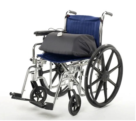 A traditional looking hospital wheelchair (dark blue, textured seat with black wheels and a shiny silver body) in in the center of a white background. On the seat rests a black assistive cushion. The cushion raises and lowers to assist individuals from a sitting to standing position more safely and easily.