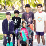 5 children, many of who appear related, gather together and look towards the camera. 4 are standing; 1 is seated in a power chair. They are all participants in the 2026 Bay Area Walk & Roll to Cure FSHD. The seated child and 2 of the standing children hold teal and orange pompoms with long strands. The child on the far right wears a 2026 Walk & Roll t-shirt.