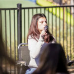 Tatiana Garcia speaks at the 2026 Bay Area Walk & Roll to Cure FSHD. Tatiana is a young, Mexican woman with long, straight, brown hair. She sits in a folding chair and holds a microphone to her mouth as she speaks to the crowd, who are out of frame. She wears a white shirt and is brightly lit by the sun.