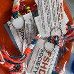A large, woven, orange bowl sits on top of a picnic table. The bowl holds an assortment of paracord keychains. The paracord designs are a collection of orange, teal, and white stripes with fringe at the end. Each cord is attached to a metal key ring, which also has an informational card attached. The message on the card thanks Walk and Roll participants for working to cure FSHD and provides information about FSHD and paracords. The keychains were created by a local company in support of a staff member who loves fishing and lives with FSHD.