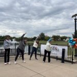 Participants at the 2026 St. Louis Walk and Roll to Cure FSHD strtch as a group before embarking on their walk around a small retaining pond. These four participants wear sweatshirts and pants and stand in front of the retaining pond, a black safety fence, and the Walk and Roll banner and a series of orange, blue, and silver balloons. The participants are standing with their legs spread ~hips width apart and are reching up over their heads with their right arms, stretching their arms, shoulders, and sides.