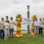 11 staff members at Avidity, who participated in the 2026 San Diego Walk and Roll to Cure FSHD stand in 2 lines, one on either side of the Walk and Roll to Cure FSHD banner. Each line starts with a small balloon tree made of green, orange, and yellow balloons. The backdrop is a gorgeous shot of a cloudy sky and the San Diego bay.
