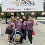 يقف ويجلس 4 أفراد يرتدون قمصان فريق عنابية اللون تحت لافتة "Walk & Roll to Cure FSHD" البيضاء. توجد على اللافتة أيضًا شعارات جمعية FSHD ومؤسسة FSHD Canada Foundation في الأسفل. يرتدي جميع الأفراد الأربعة تعبيرات وجه سخيفة مع خروج ألسنتهم. الرجل الذي في المنتصف يجلس على سكوتر كهربائي والنساء الثلاث واقفات جميعاً.
