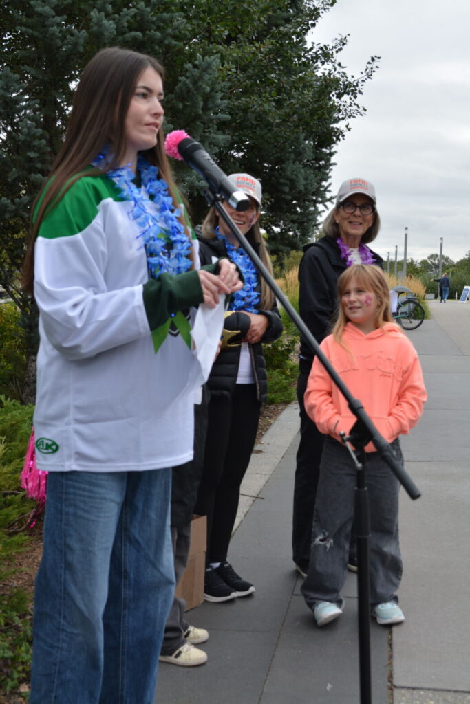 Quinn Wilson, a young woman living with FSHD stands speaking into a microphone. Quinn has long, straight honey brown hair and is wearing blue jeans and a white and green windbreaker. She is addressing a crowd; the crowd is outside the frame. Near her stand a few event participants in cold weather gear.