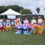 7 participants at the 2026 San Diego Walk and Roll to Cure FSHD stand wearing an assorted collection of air-inflated costumed. The costumes stop at their midsections. 5 are brightly colored chickens and 2 are blue snails with pink shells. Apparently, you had to be there to truly understand the whimsy and fun depicted in this photo.