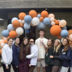 7 young women stand around 1 young man underneath a balloon arch. The balloons are a mix of large and small balloon in white, orange, and light blue. The young man is taller than everyone, wears glasses and a 2026 Walk & Roll t-shirt and is holding up a peace sign. Everyone looks towards the camera with hopeful, happy expressions.