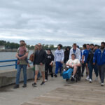 Participants at the 2026 Michigan Walk & Roll to Cure FSHD walk and roll along a concrete path at the water's edge. They wear jackets and sunglasses. One individual is in a wheelchair with a knee brace on.